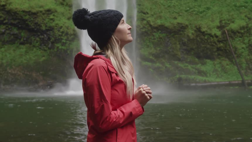 A young woman looking down and praying then looking up to the sky with a waterfall and lake behind her