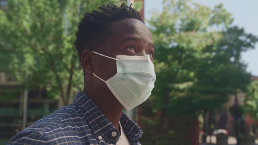 A man wearing a medical mask raises his head to look up at the sky while standing outdoors in a city during the day