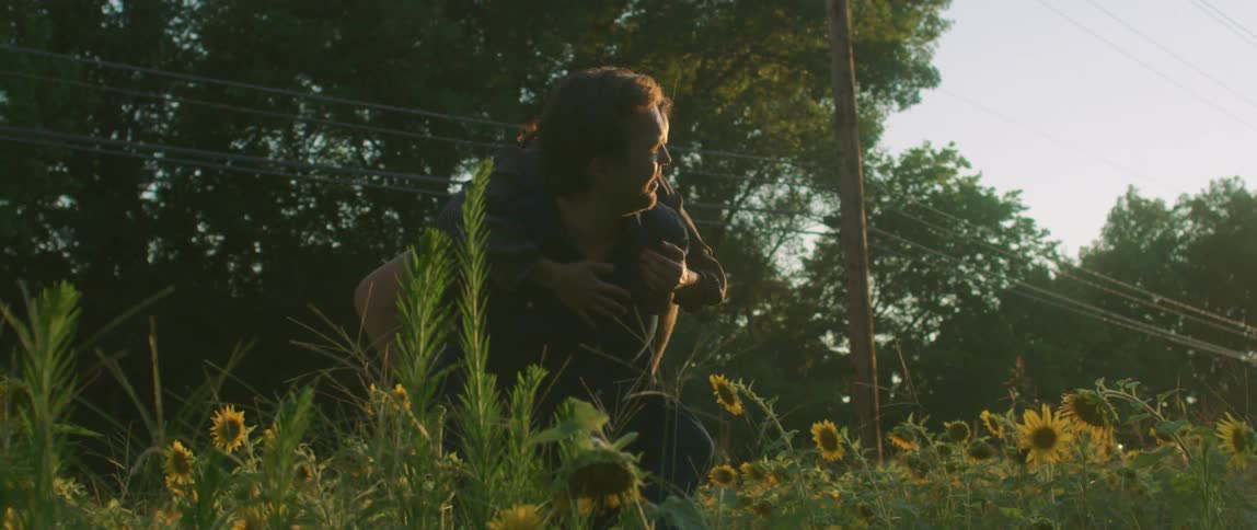 A couple stand in a field of sunflowers and the woman jumps on her husbands back and they laugh