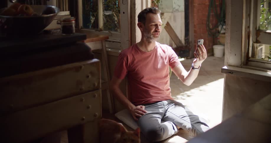 A man in a room sitting on the floor talking on a smart phone next to a cat