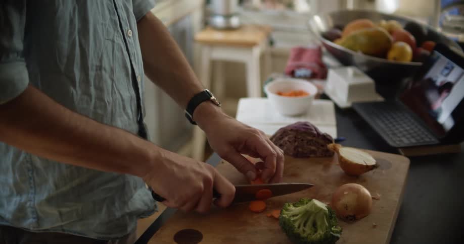A man cuts produce in a home kitchen while having a video call on a tablet