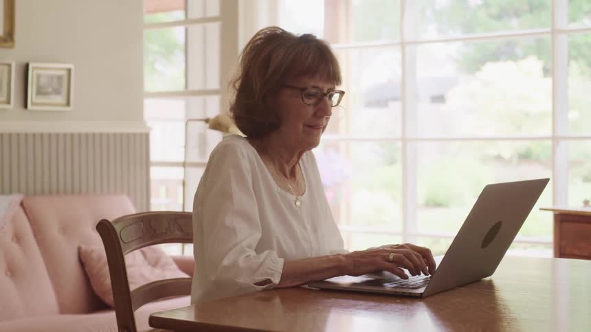Older woman sitting at dining room table typing on a laptop