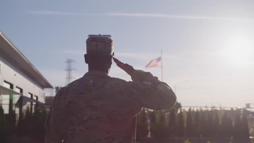 A man in a military uniform standing and saluting the american flag with trees the sky and clouds in the background