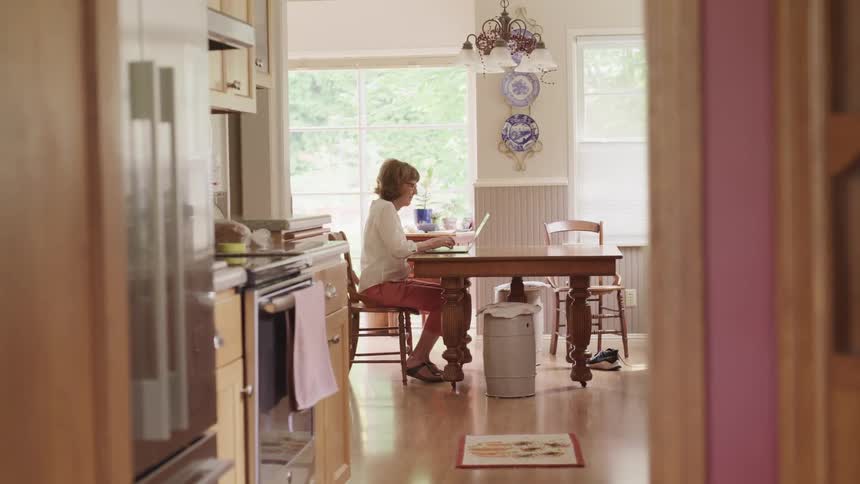 Older woman sitting at table in kitchen typing on a laptop