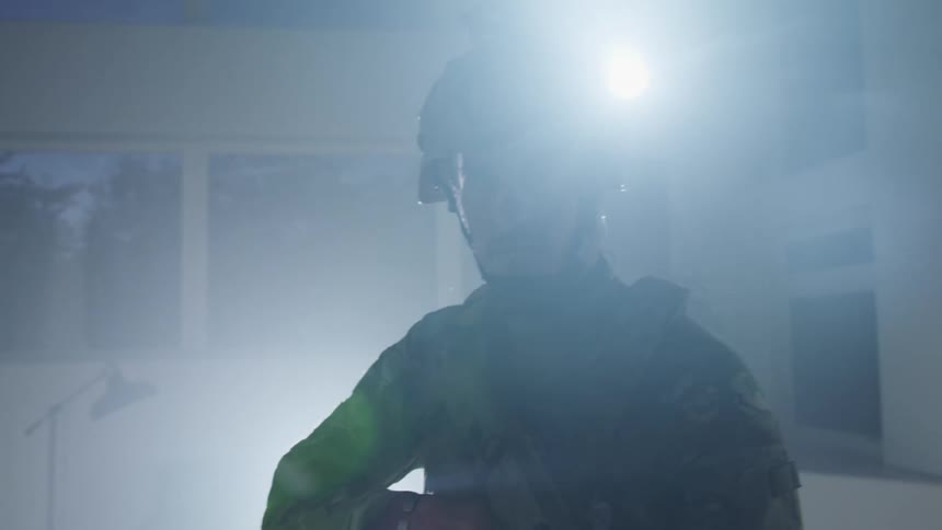 An armed soldier in combat uniform stares into the camera while standing in a dark room at night