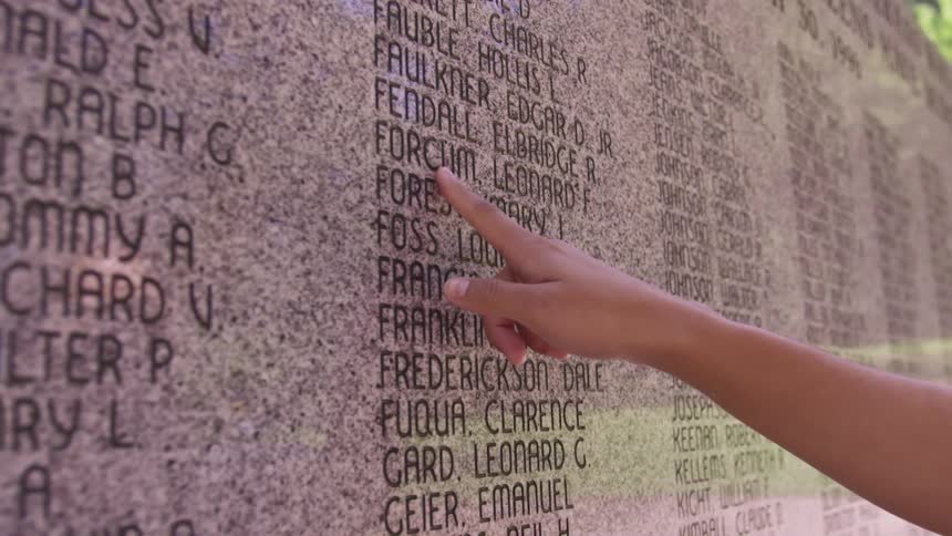 A person by a memorial pointing out names with trees the sky and clouds in the background