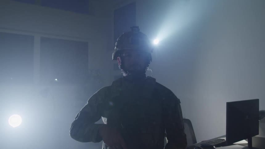 A soldier in tactical gear including light on his helmet and carrying a weapon stands in a room near a desk
