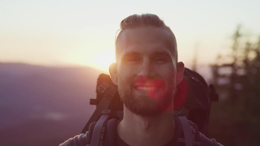 A young man smiling wearing a hiking backpack with forest and mountains in the background