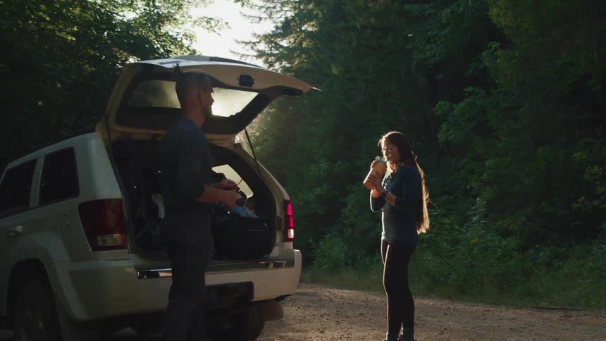 A man puts hi sunglasses up on his head as his wife takes a sip from her drink as they both stand near the back of their suv loaded with hiking equipment