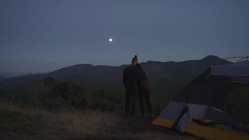 A man and woman embrace while looking out over a forested countryside from a hill at night