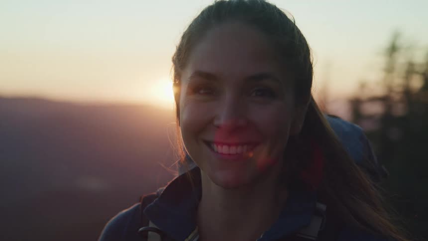 A young female hiker smiles into the camera while standing outdoors at sunrise