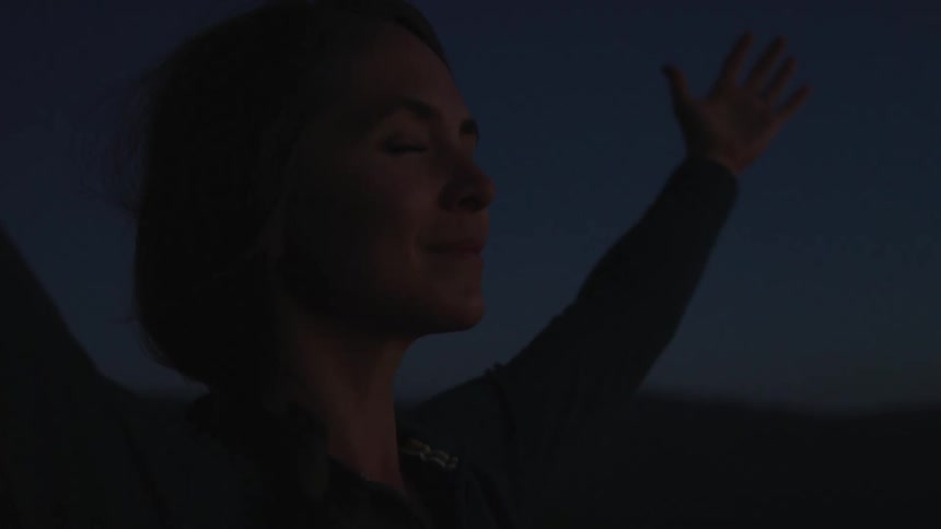 A woman closes her eyes while standing outdoors with her arms held wide at night