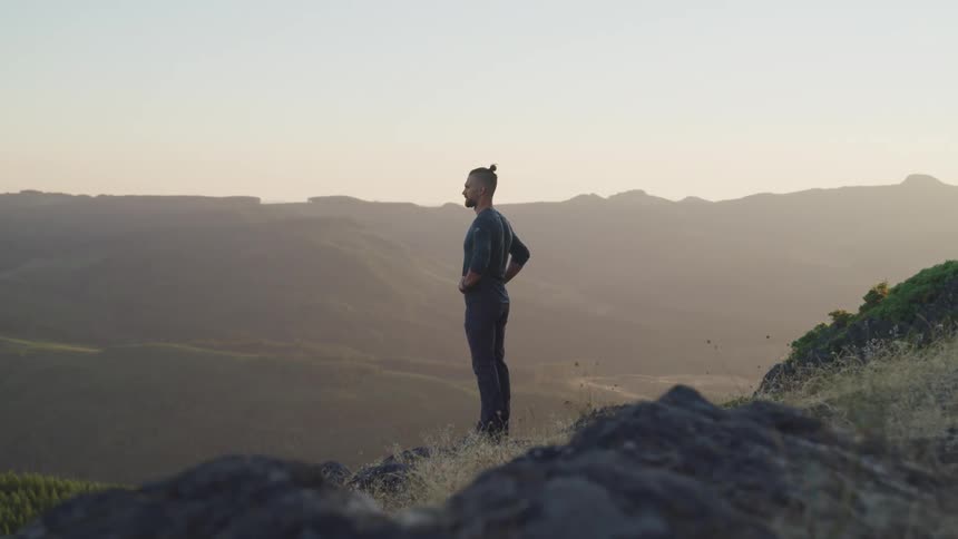 Young man standing still on top of a mountain looking at the hills at sunset