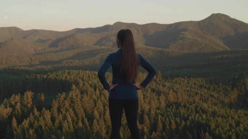A woman standing outside with trees a mountain range the sky and clouds in the background