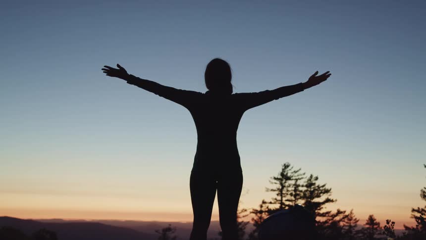 A woman raises her arms out and up as she watches the sun set in a canyon