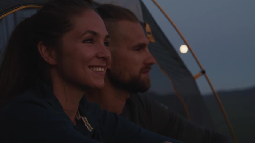 A man and woman sit at the edge of their tent while enjoying the evening sky