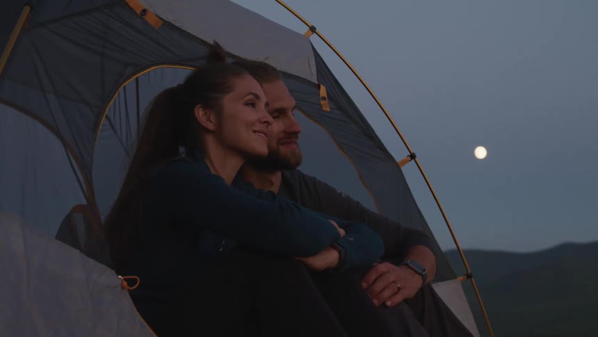 A married couple sitting close together in a tent looking out and smiling at dusk