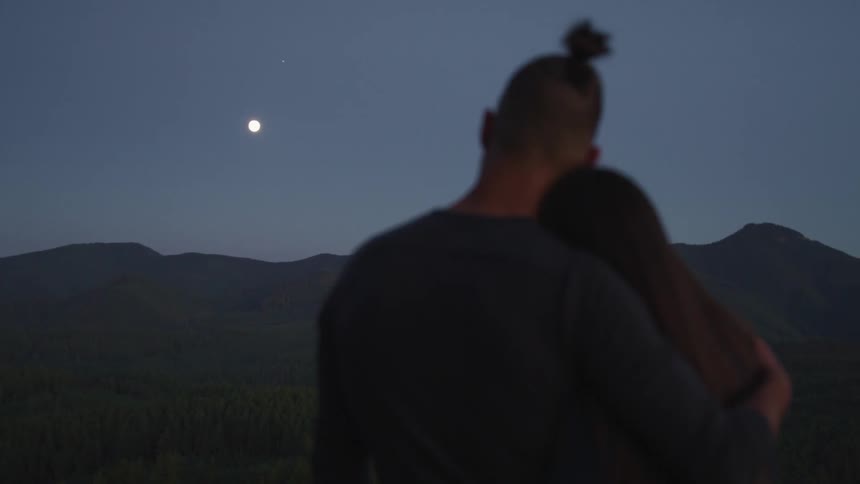 A man and woman embrace while looking out over a forested countryside at night