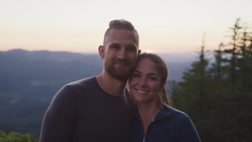 A man and woman stand close and smile with trees and mountain behind