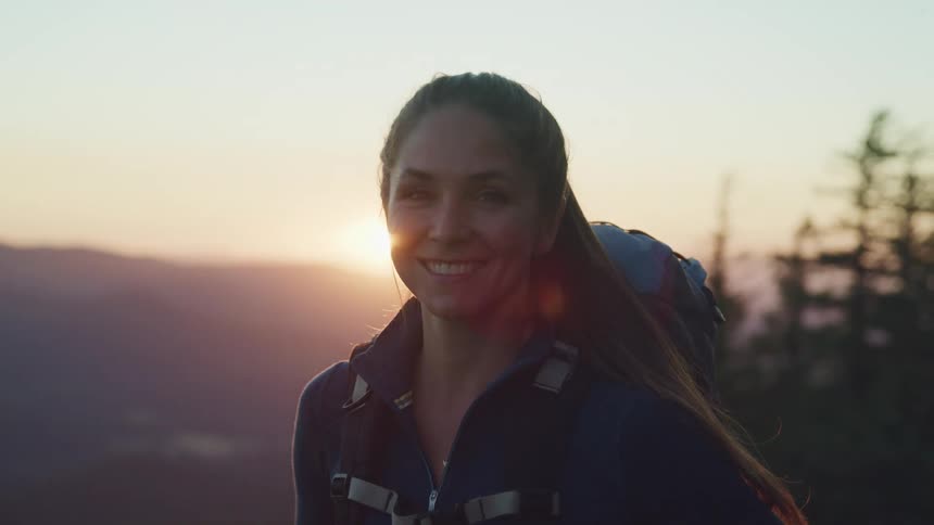 A woman with a backpack standing staring and smiling with trees the sky and clouds in the background