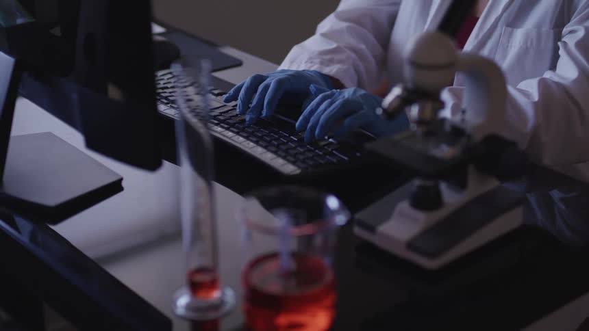 A scientist wearing disposable gloves sitting a desk typing with a microscope and beakers beside them