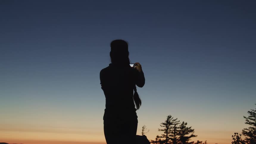 Silhouette of a young woman filming the sunset over the mountains with a camera