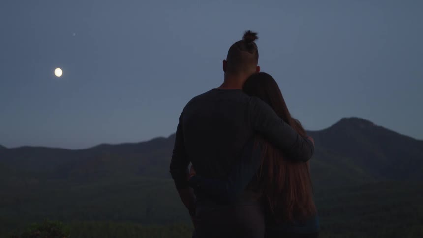 Young couple hugging while looking at the moon over the mountains at evening
