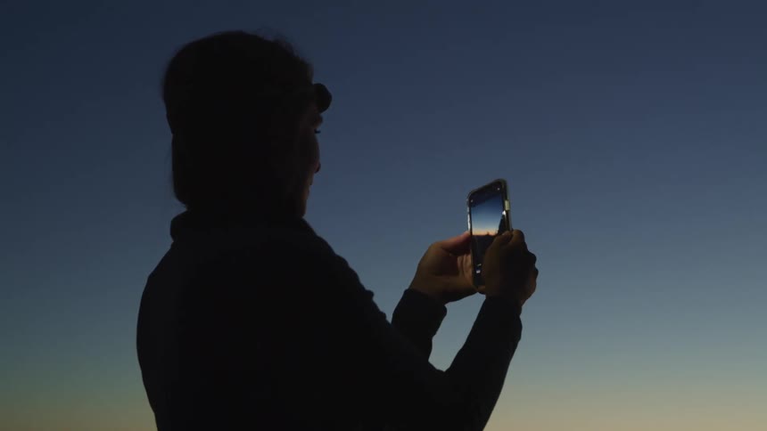 A person standing outside taking a picture on a smart phone with the sky and clouds in the background