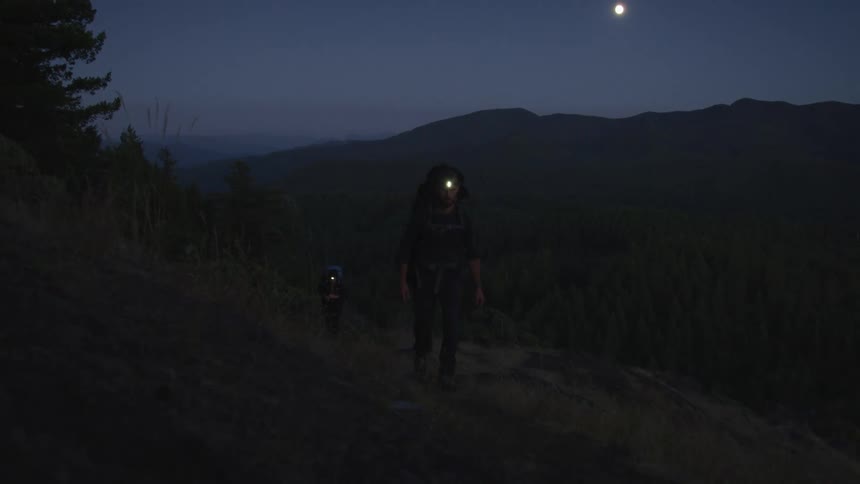 A male and female hiker hike along a mountain slope overlooking a vast forest at night