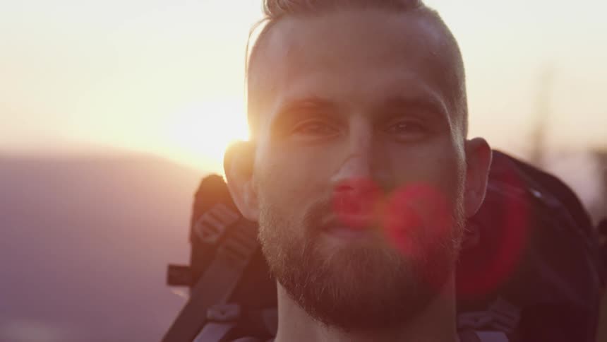 Young bearded man wearing a backpack looking at the camera while the sun sets behind him over the mountains