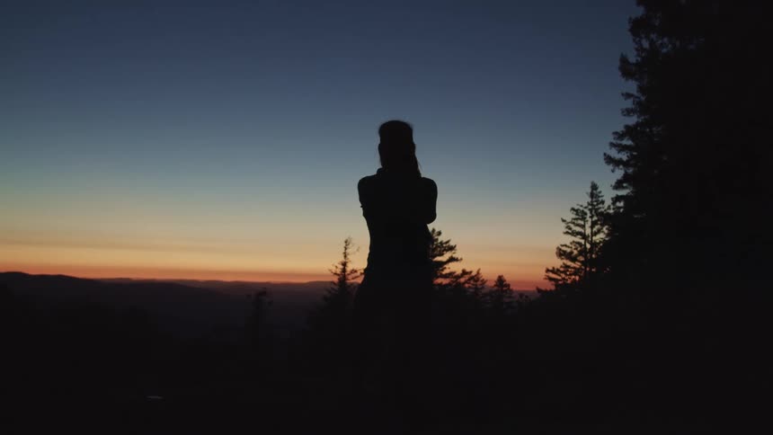 Person stands outdoors in the dark near conifer trees filming red sky with mobile phone