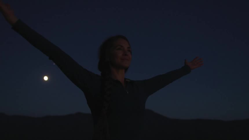 The moon shines behind a woman raising her hands up to the night sky standing outdoors