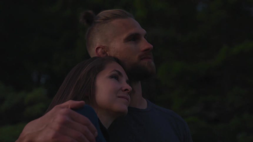 A man and a woman hugging with the man kissing the woman on her forehead with trees in the background