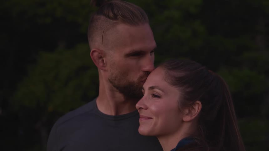 A man turns to kiss the forehead of the woman leaning against his shoulder as they stand outdoors during the day
