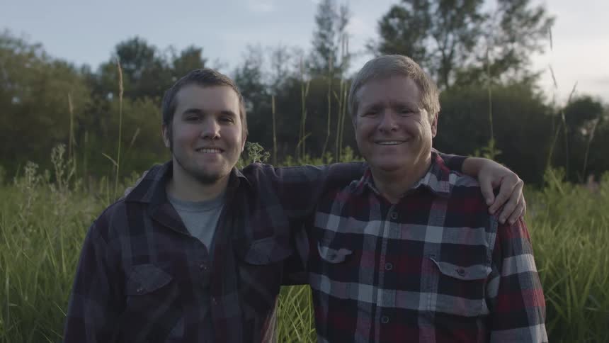 A smiling father and son posing outside with their arms around each other