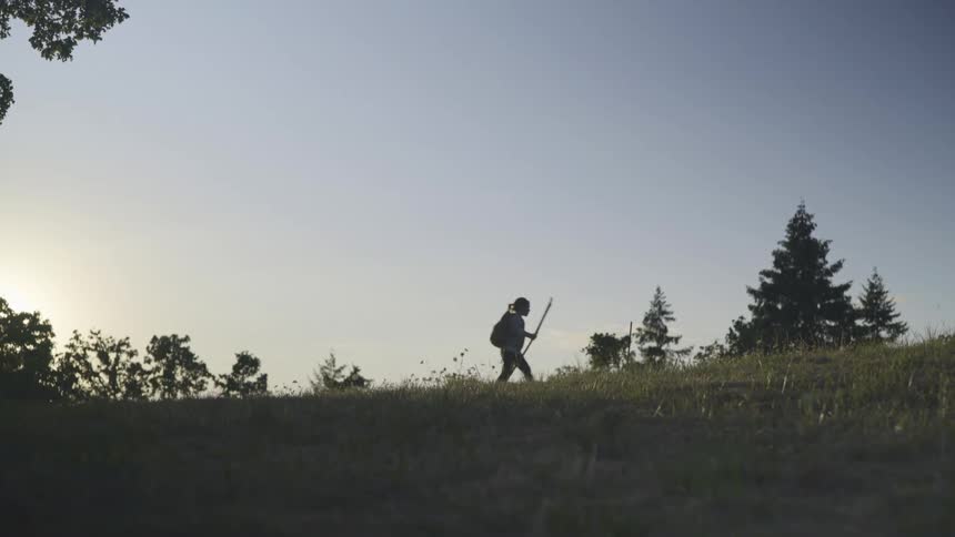 Young girl wearing a backpack uses a staff to walk on a trail over a hill in the morning