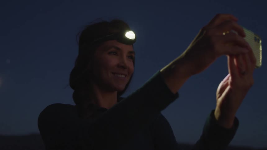 A woman with a flashlight strapped to her head looking at a smart phone with the moon and the sky in the background