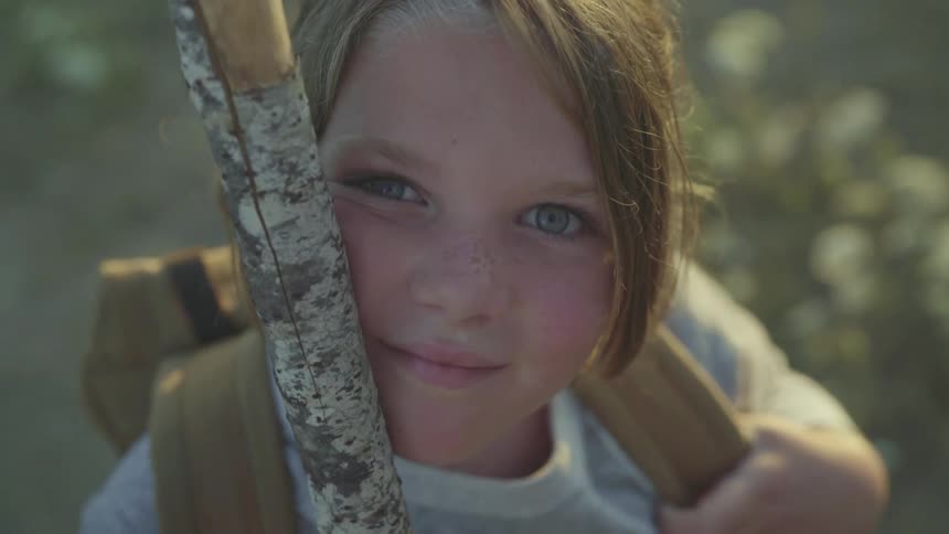 A girl with a backpack outside holding a stick with plants in the background
