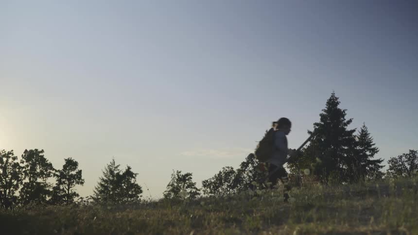 Young girl wearing a backpack is carrying a walking stick and walking across the grass with trees in the distance