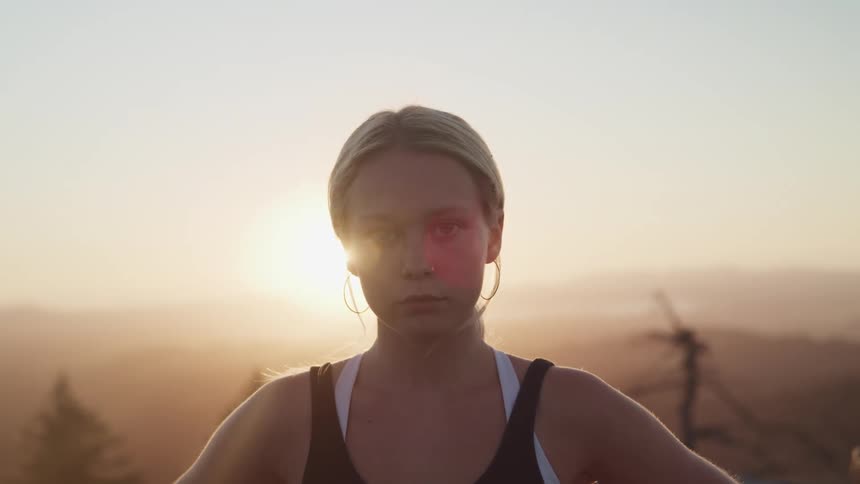 A woman stands on a hilltop with the sun shining behind her