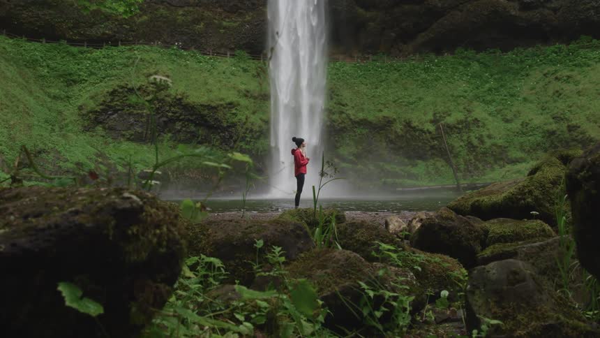 A woman with a jacket and a hat standing with a waterfall in the background