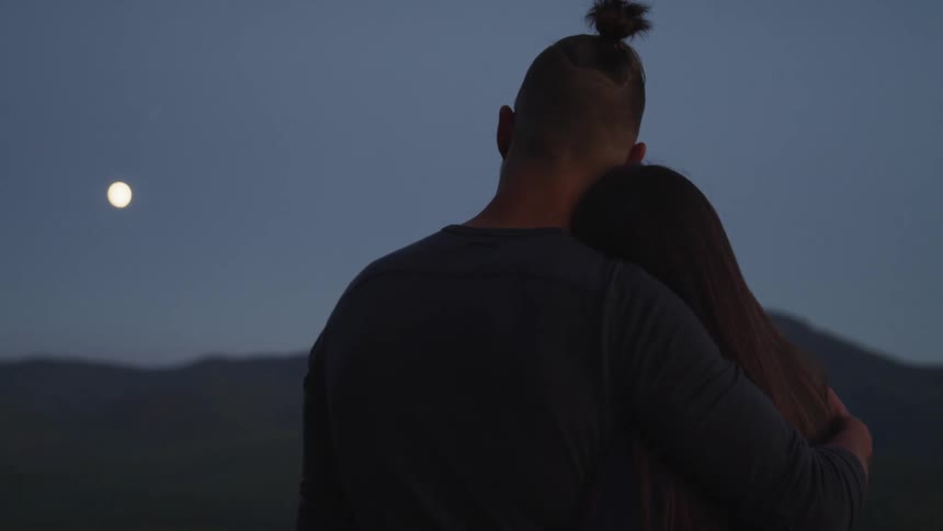 Couple affectionately holding each other watches moon at dusk