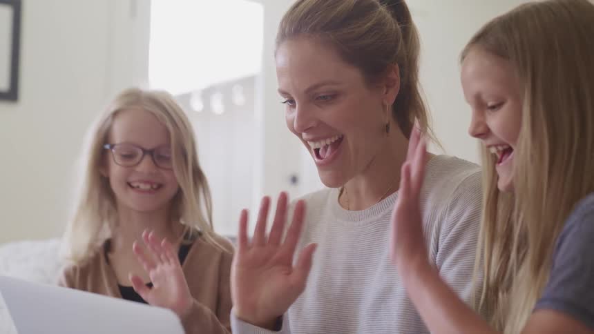A mother and her daughters smile and wave to a video call on a laptop