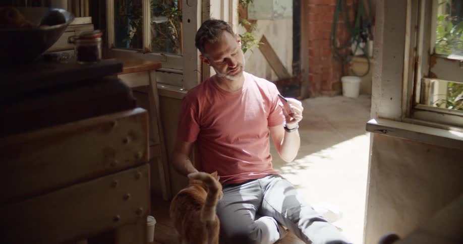 A man in a room sitting on the floor video chatting on a smart phone while petting a cat