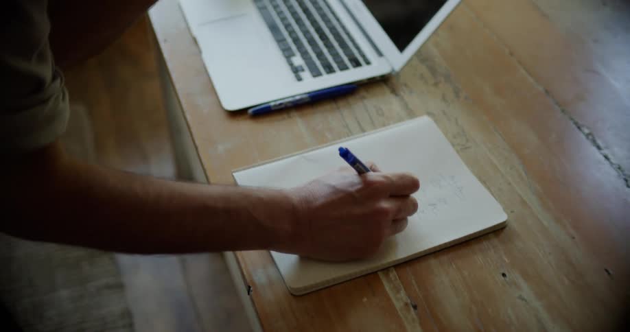 Man working from home talking on a smartphone takes notes with a pen on a notebook next to a laptop