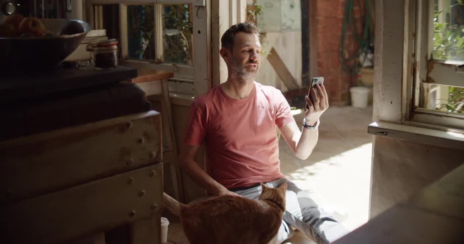 A man in a room sitting on the floor video chatting on a smart phone while petting a cat