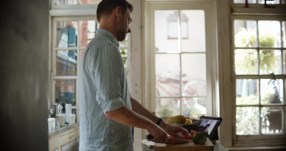 A man stands and cuts food on a cutting board while carrying on a video conversation via his tablet computer