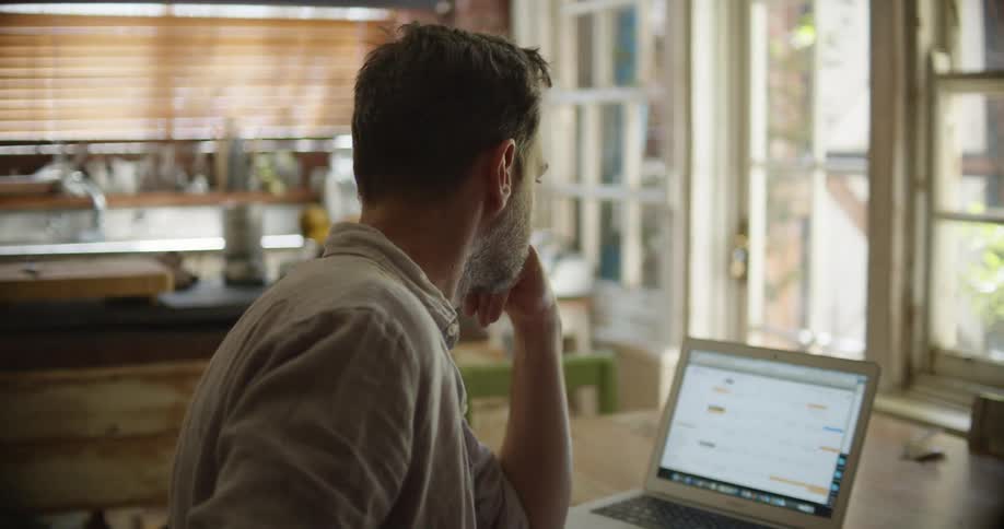A man in a room sitting at a desk with a laptop writing on paper with a pen