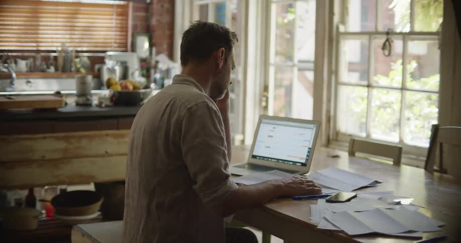 A man sits at a table at home with his laptop and cell phone and goes through paperwork
