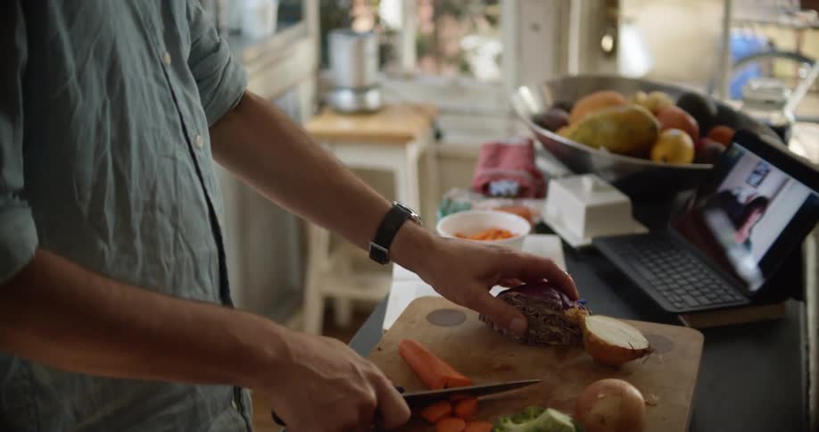 A man chops produce with a large knife while having a video call on a tablet